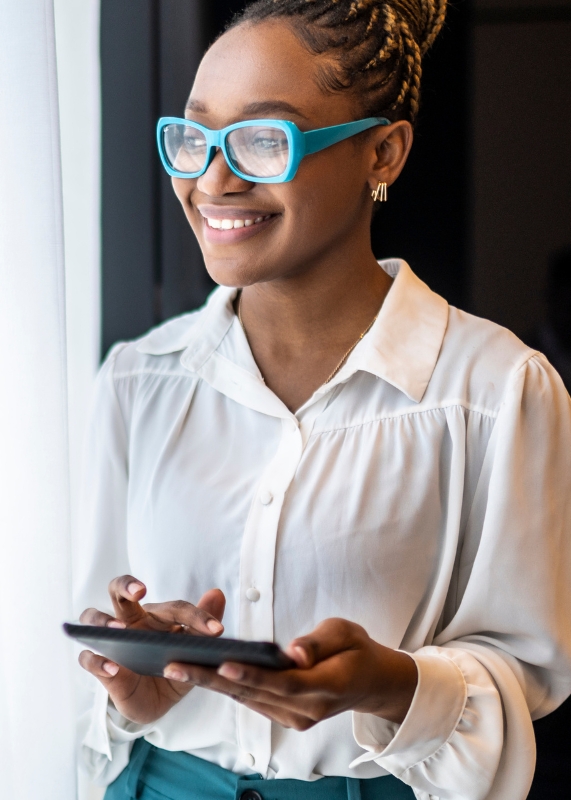 A photograph of a Black Woman wearing glasses looking at something while holding a smartphone.